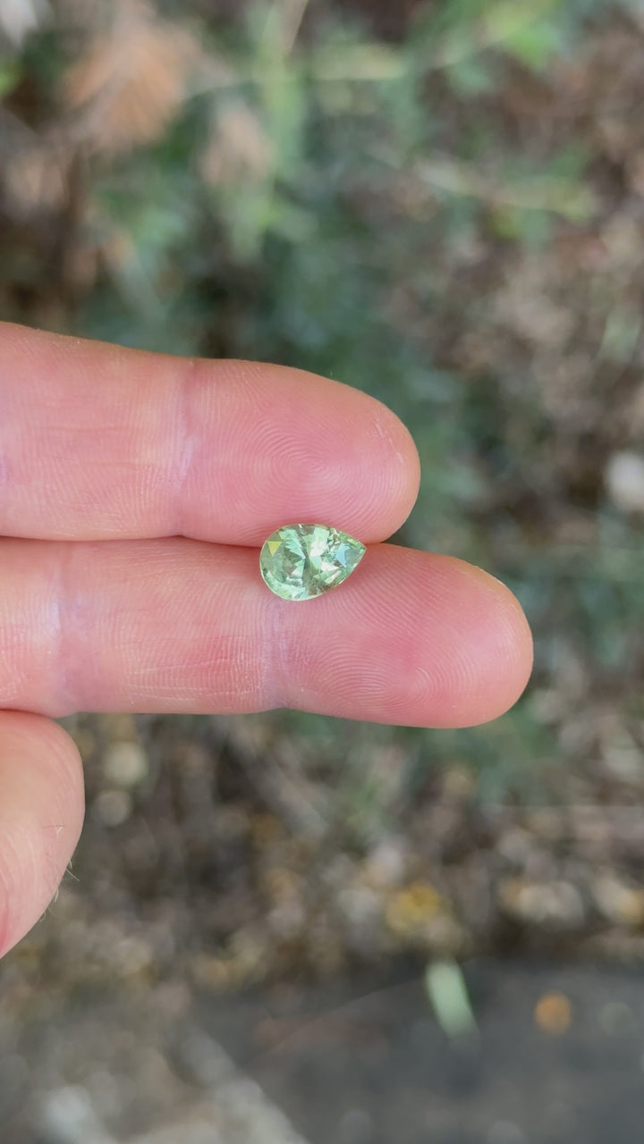 Rotating view of a 3.04ct pear-shaped sage green and peach sapphire held in fingers under natural daylight, showcasing its brilliance and color play