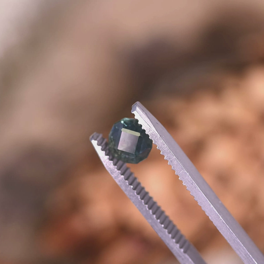 ideo of 1.32ct Montana sapphire in tweezers rotating under light, blue green teal geometric rounded cushion cut showing natural brilliance and facet reflections