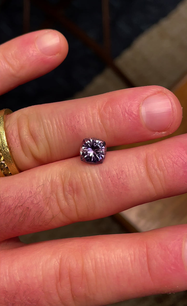 A 3.13-carat cushion-cut color-change sapphire held between fingertips, showcasing its grayish-green to pinkish-purple hues from Montana, USA.