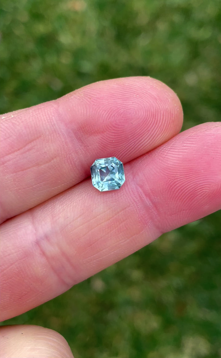 A high-definition video showing a 1.59-carat opalescent powder teal sapphire held in tweezers, highlighting its octagonal step cut and brilliance in natural light.