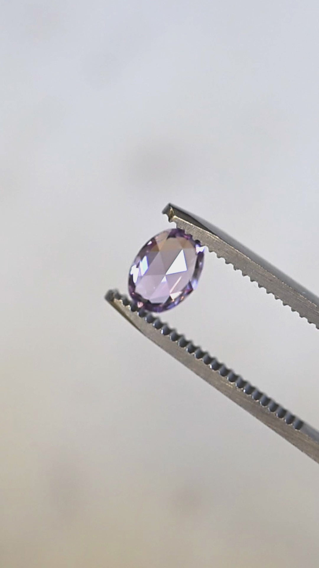 Natural Madagascar sapphire turning in tweezers, showcasing violet, lavender, and pink color play with a luminous opalescent glow.