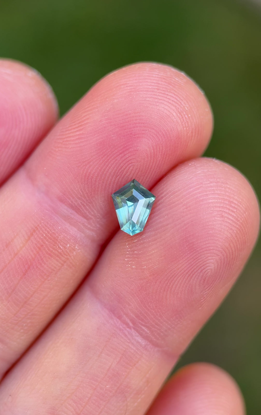 Close-up hand rotation video of a 1.14ct custom shield cut parti sapphire from Montana, highlighting its vivid teal and green tones and light performance against a natural outdoor background.