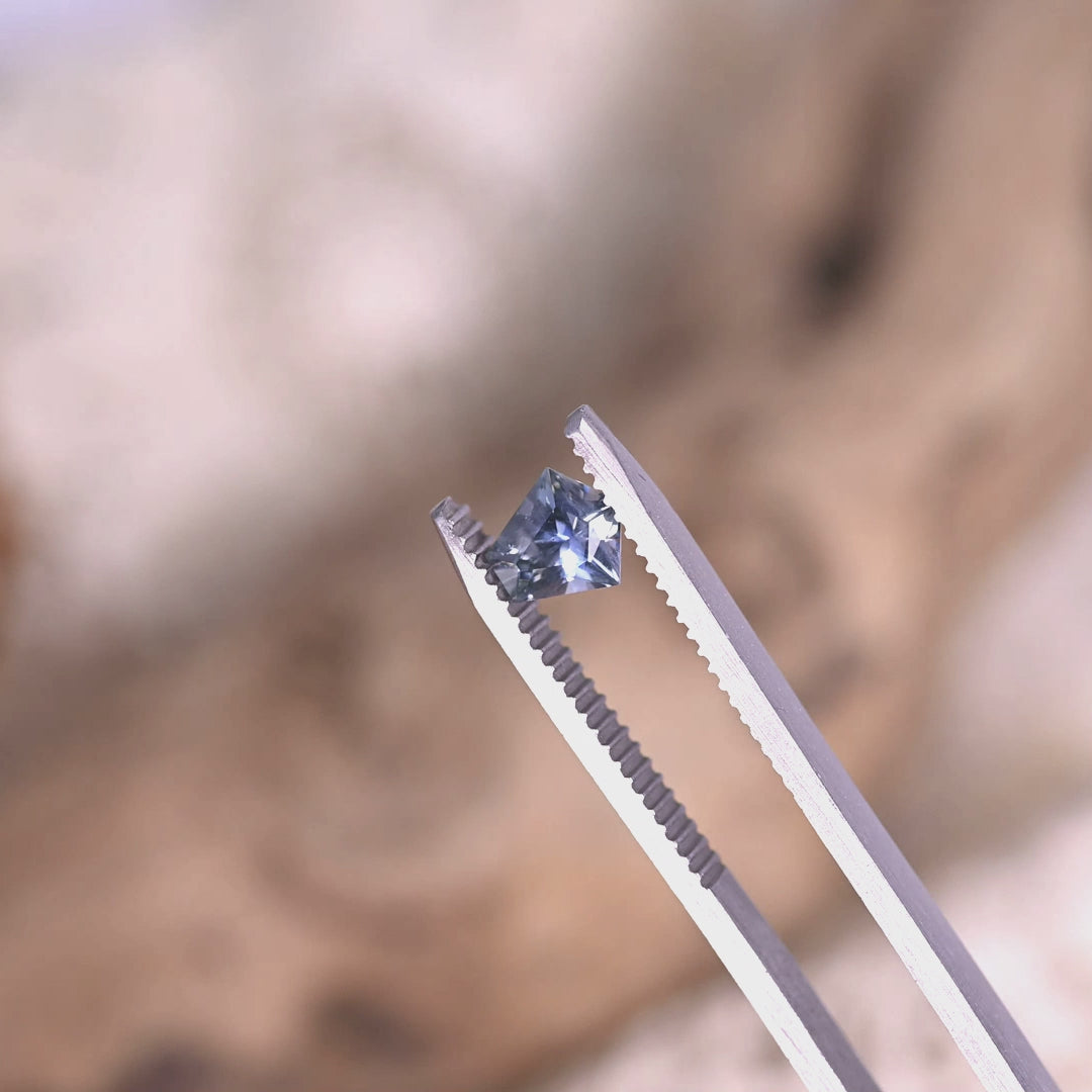 0.85ct Montana sapphire modified kite cut rotating in tweezers under natural light, displaying blue, green and violet color shift for custom fine jewelry.