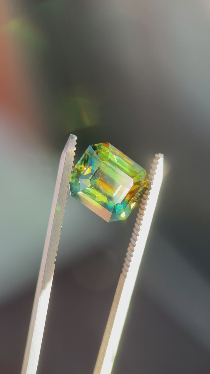 Slow-motion video of a 9.14ct Nigerian parti sapphire in a precision emerald cut, highlighting broad step facets, transparency, and vivid orange, yellow, blue, and green color zoning from Gombe, Nigeria