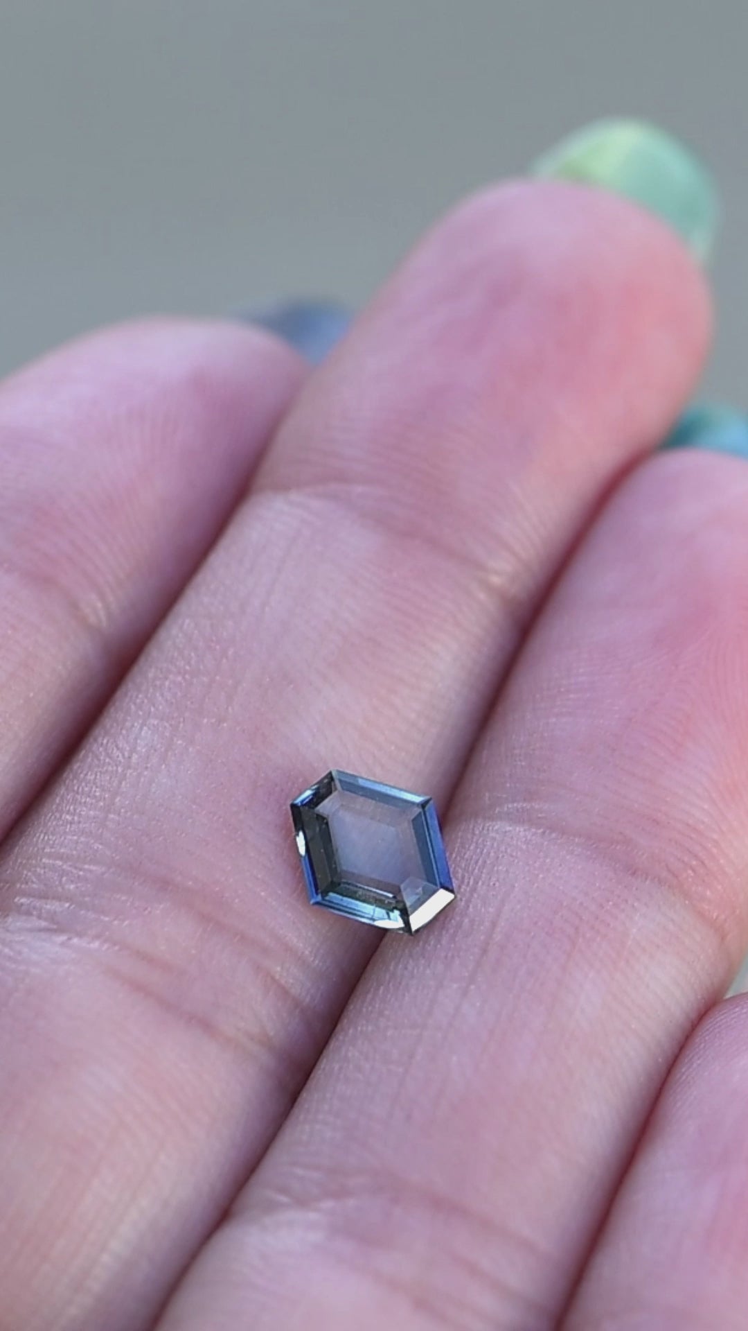 Hexagonal portrait cut teal blue 1.02ct sapphire resting and rotating on fingertips under natural light.