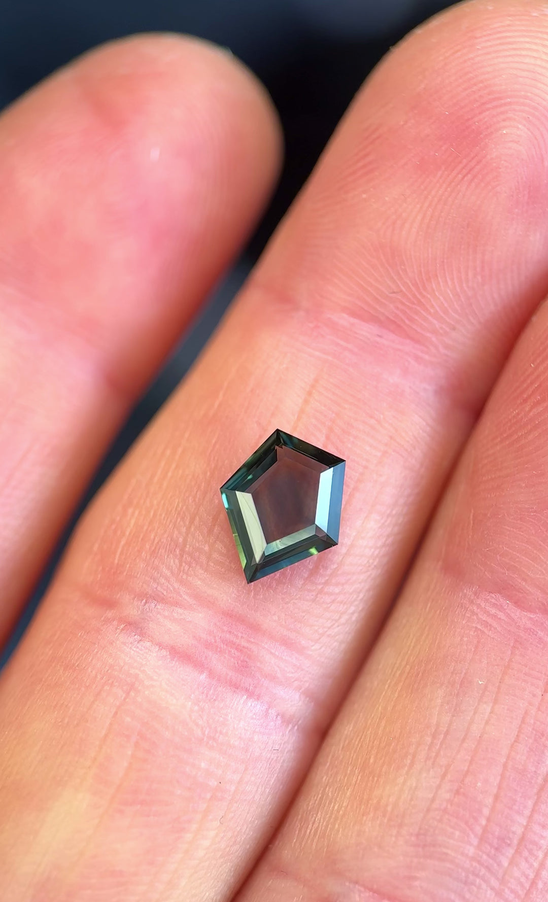 Loose custom shield portrait cut sapphire turning in tweezers, displaying natural blue and teal tones from Gombe, Nigeria.