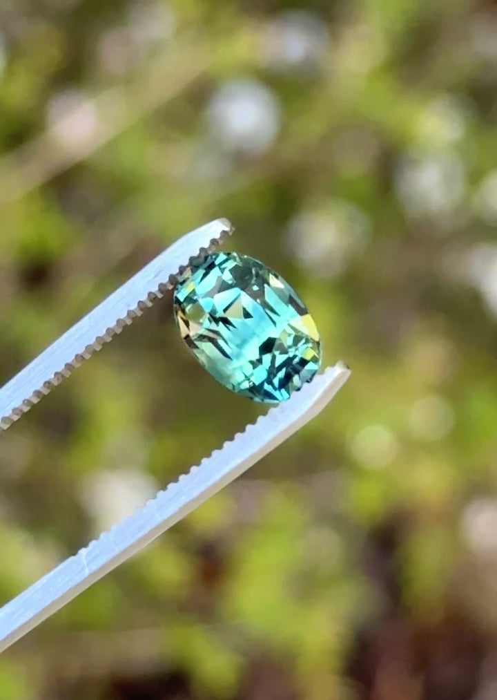 Natural Madagascar parti sapphire turning in tweezers, showcasing shifting green, teal, and blue tones with lively brilliance.