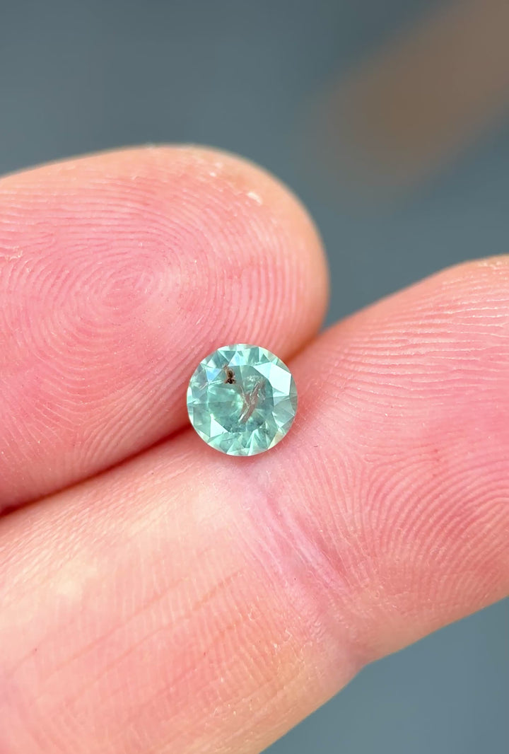 1.10ct opalescent powder teal sapphire rotating in hand, revealing soft seafoam blue flashes and round cut brilliance.