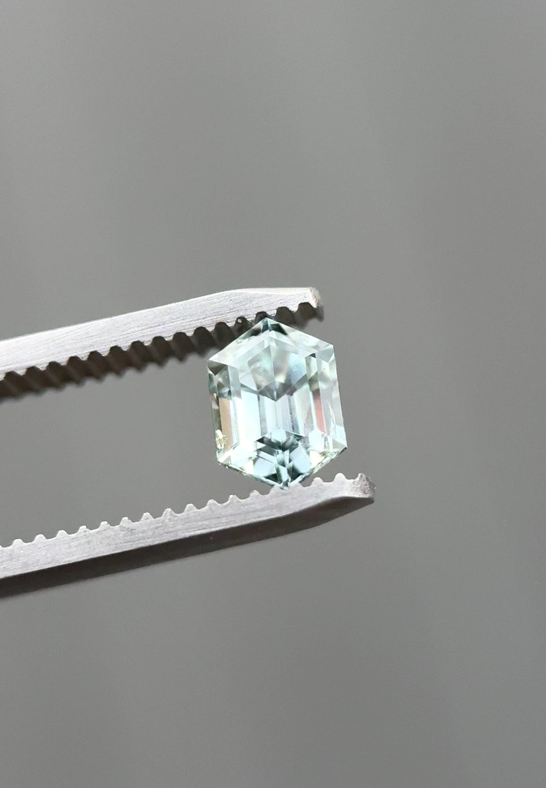 Video of a 1.22 carat Montana sapphire slowly rotated in jeweler’s tweezers, revealing precision elongated hexagon step cut facets and a natural color shift from light seafoam green to grey silver under changing light.