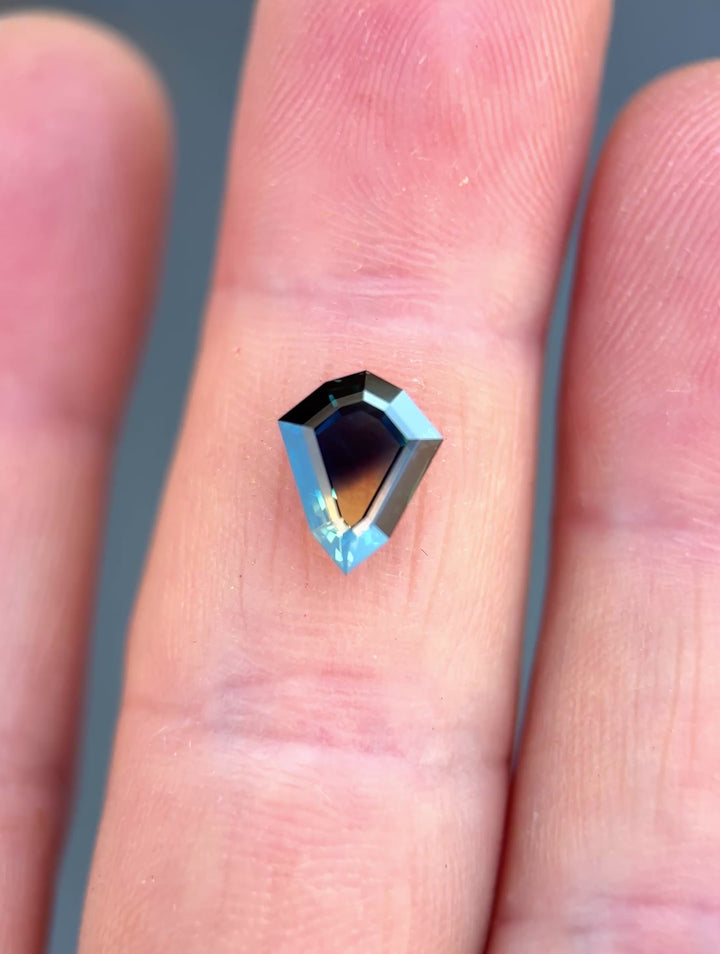 1.87ct Australian parti sapphire rotating in hand, revealing shifting yellow, teal, and blue color zones across the custom shield portrait cut.
