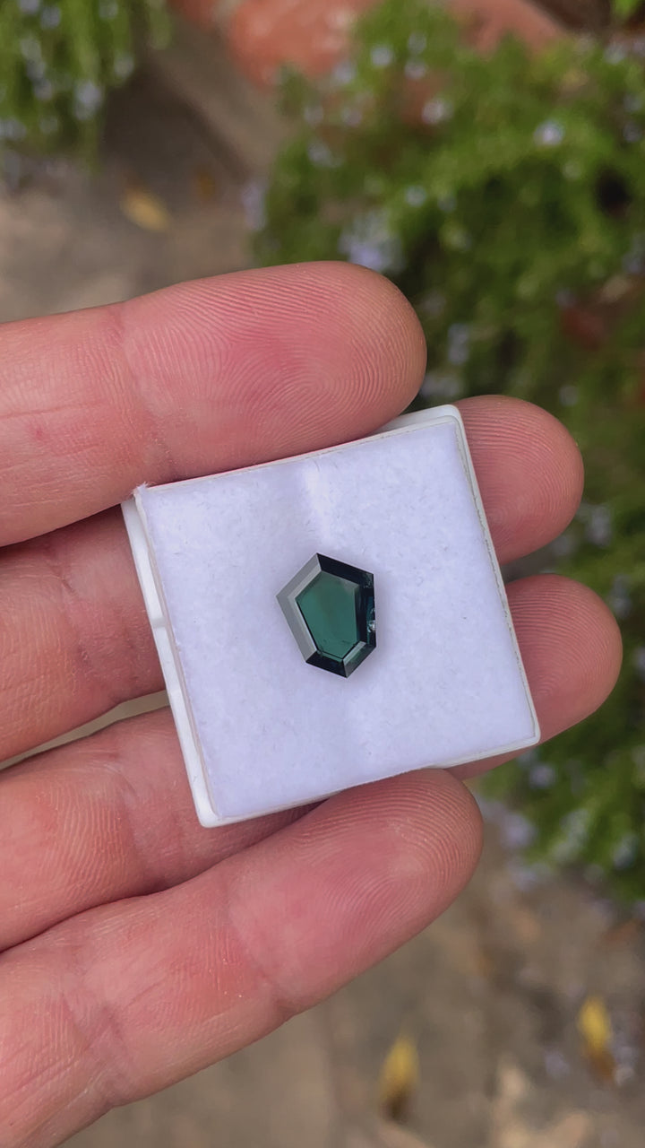 A rotating 3.77-carat sapphire with a custom shield portrait cut shows off its deep peacock, teal, blue, and green hues. The video captures the gemstone’s vivid colors and clean facets as it rotates under natural light. The untreated stone from Garba Tula, Kenya, measures 12.54mm by 9.29mm by 3.02mm.