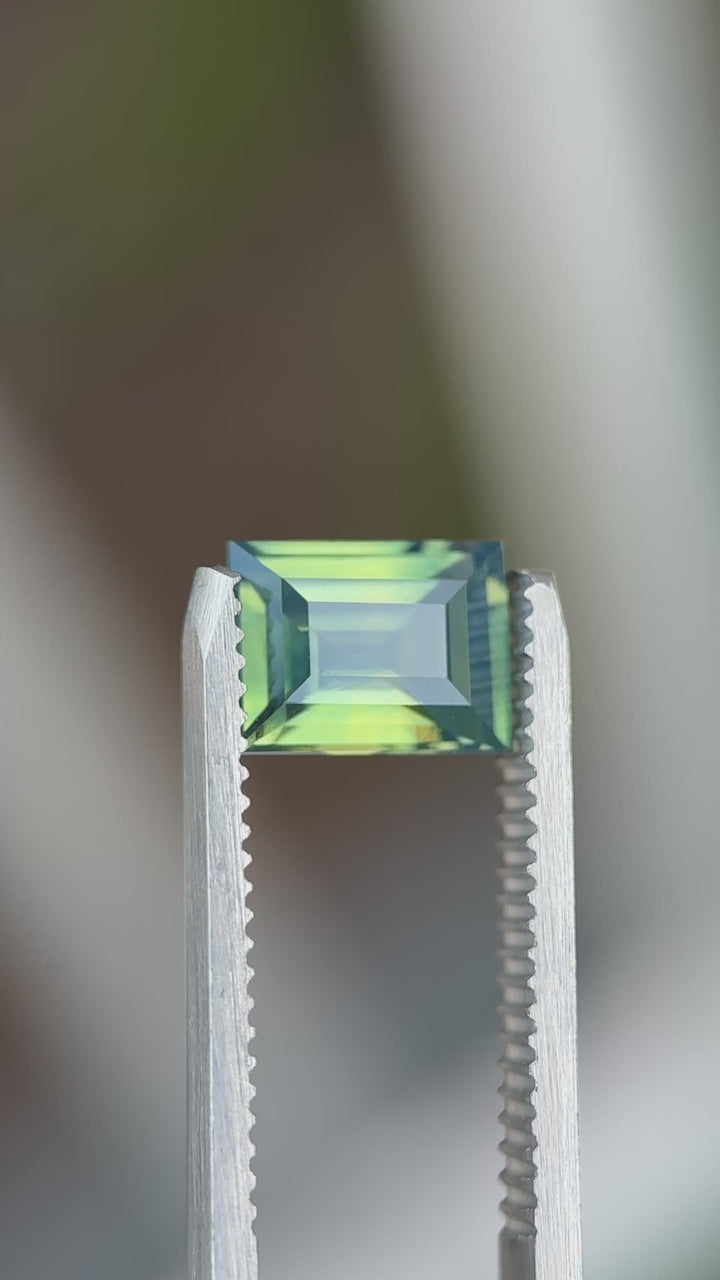 Natural Madagascar sapphire turning in tweezers, showcasing baguette step faceting and subtle teal-to-blue color movement.