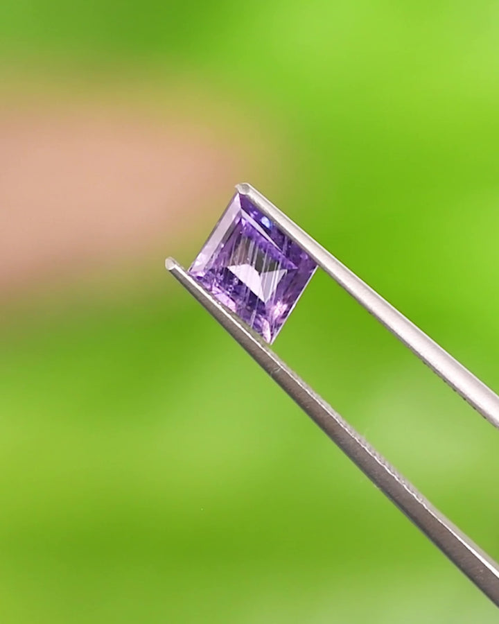 Video of 1.40CT purple sapphire rotating in gemstone tweezers, showing light reflections, clarity, and detailed step cut facets of the rhombus-shaped, unheated sapphire from Umba River Valley, Tanzania.