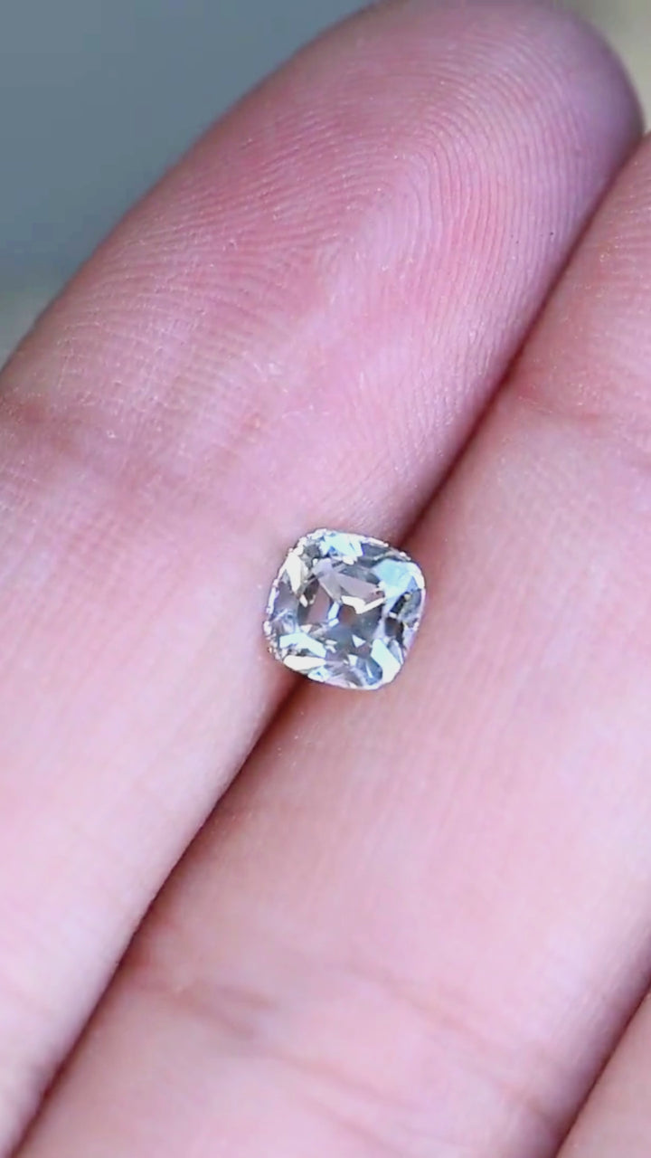 A 0.79 carat white antique-style cushion cut diamond rotating on fingertips in natural light, revealing broad flashes of brilliance and a timeless vintage-inspired facet pattern.