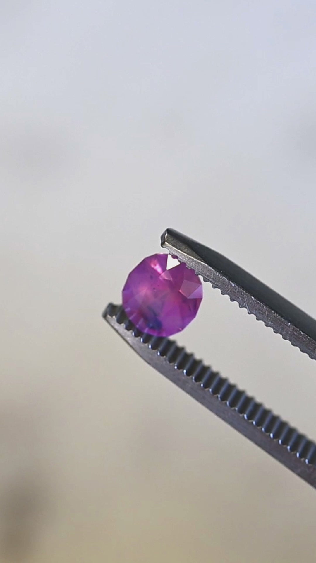 Natural Sri Lankan pink sapphire turning in tweezers under light, showcasing oval mix cut faceting, opalescent glow, and rare blue zoning.
