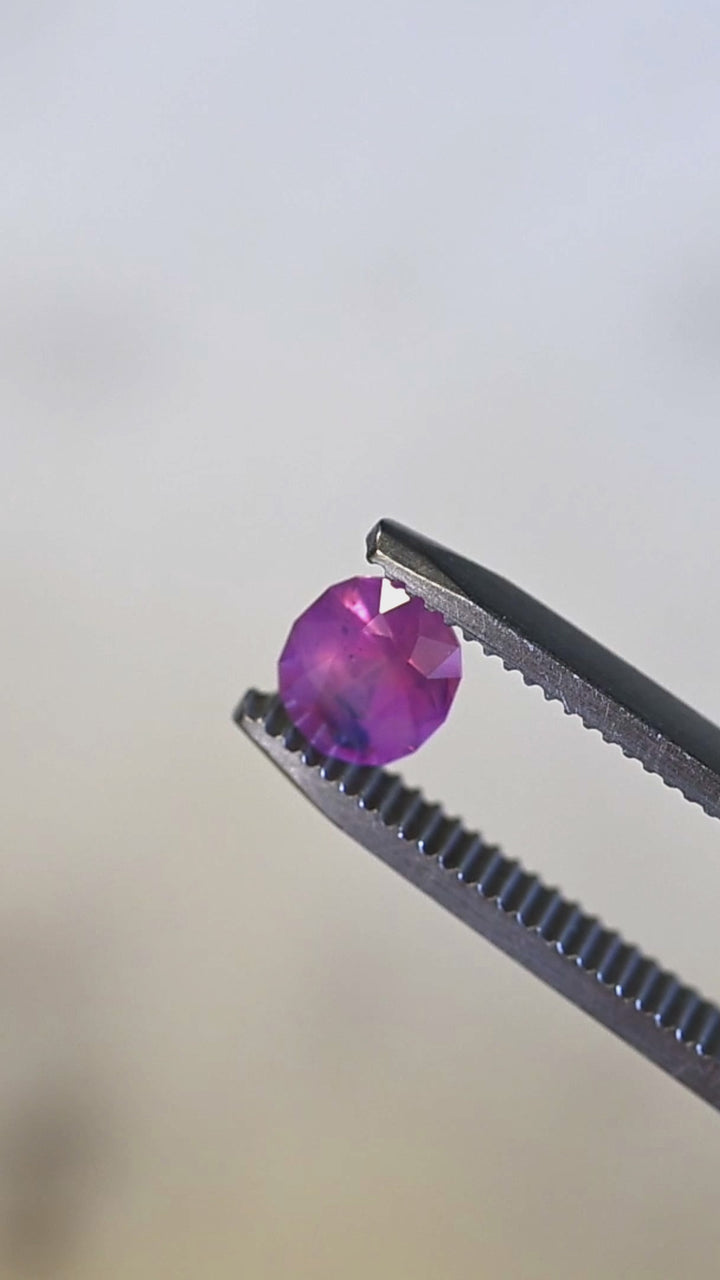 Natural Sri Lankan pink sapphire turning in tweezers under light, showcasing oval mix cut faceting, opalescent glow, and rare blue zoning.