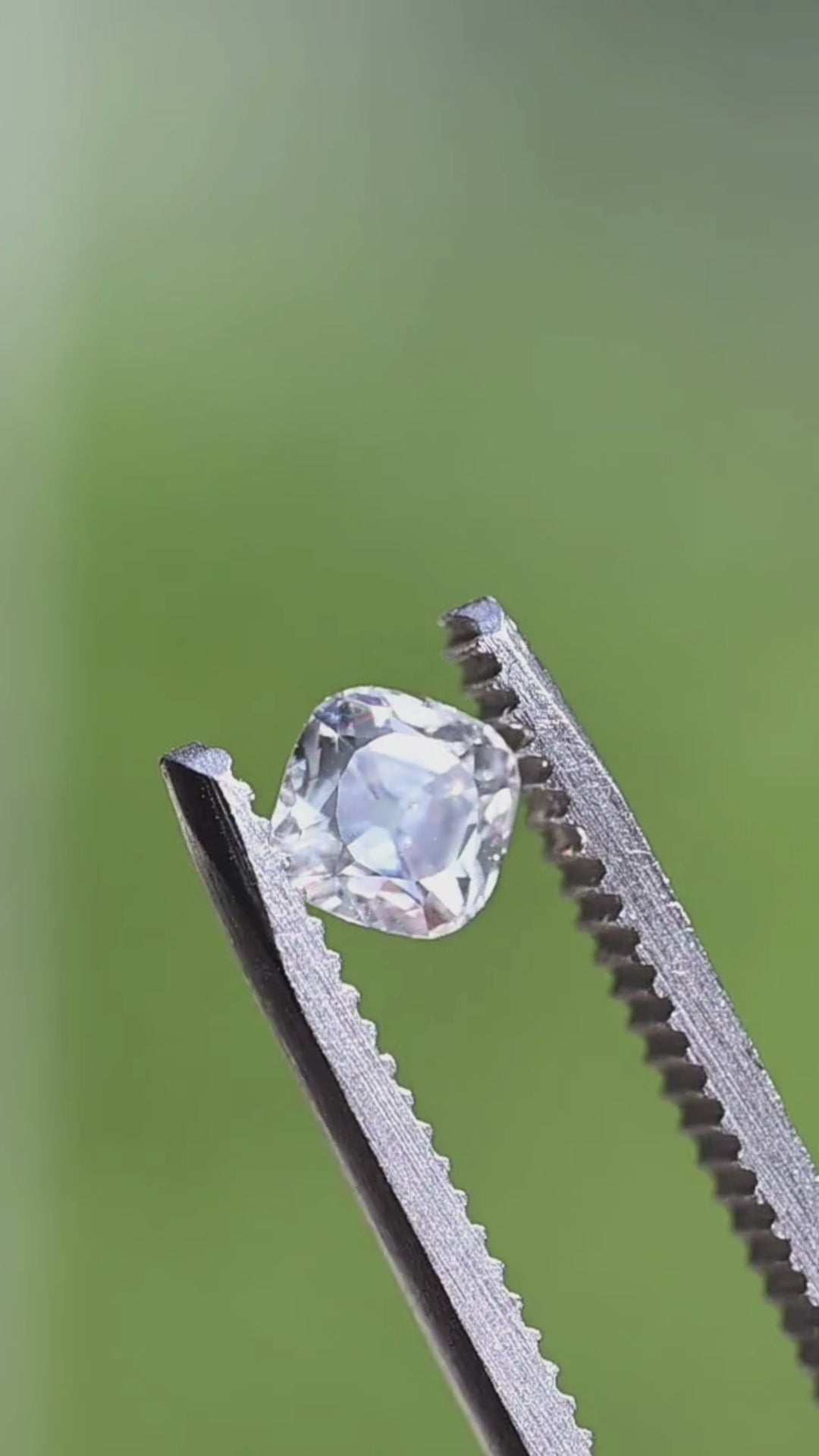White antique-style cushion diamond slowly turning in jeweler's tweezers, highlighting its balanced proportions, bold reflections, and elegant Old Mine inspired character.