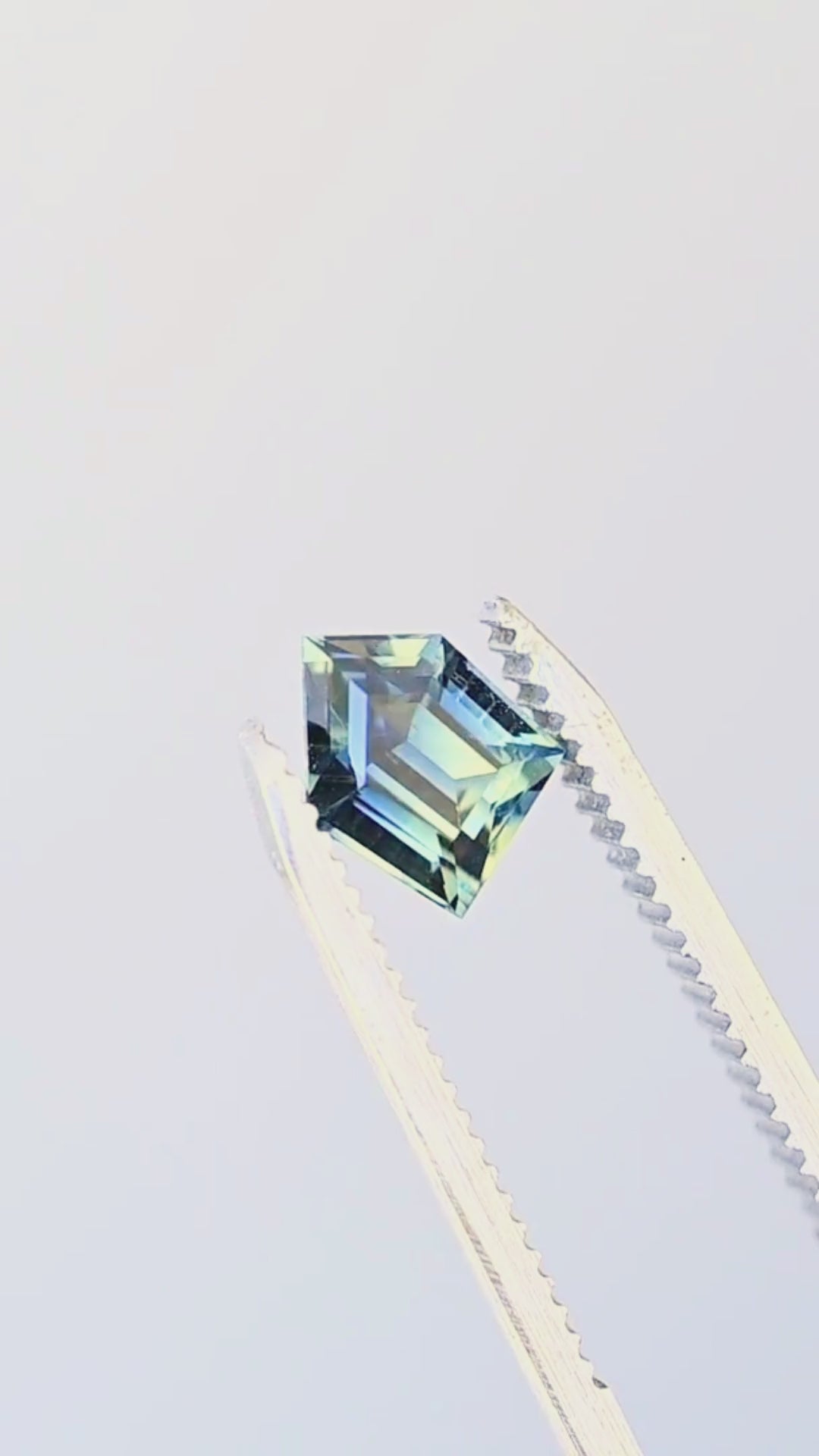 Freeform parti sapphire rotating in jeweler’s tweezers, showing its angular profile, broad step-cut reflections and shifting natural zones of blue, teal, green and yellow.