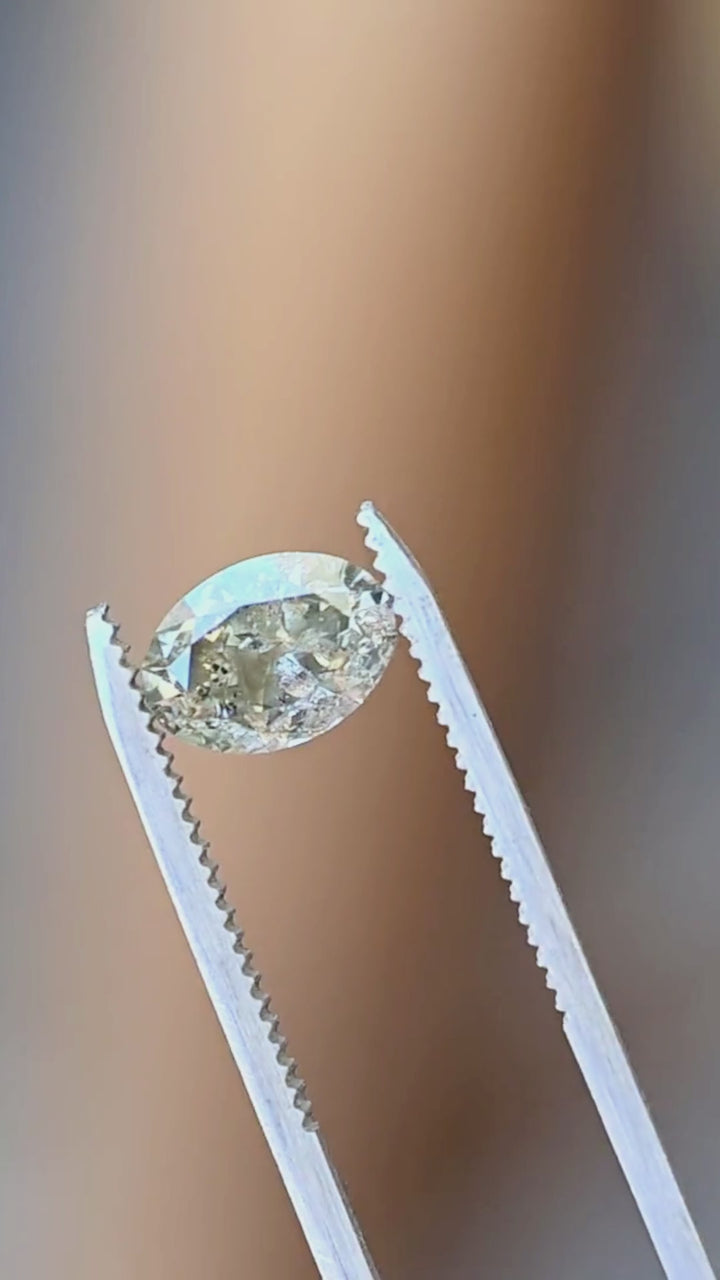 Fancy greenish brown grey salt and pepper oval diamond held in metal tweezers against a pale background, showing its brilliant faceting, elongated proportions and included interior.