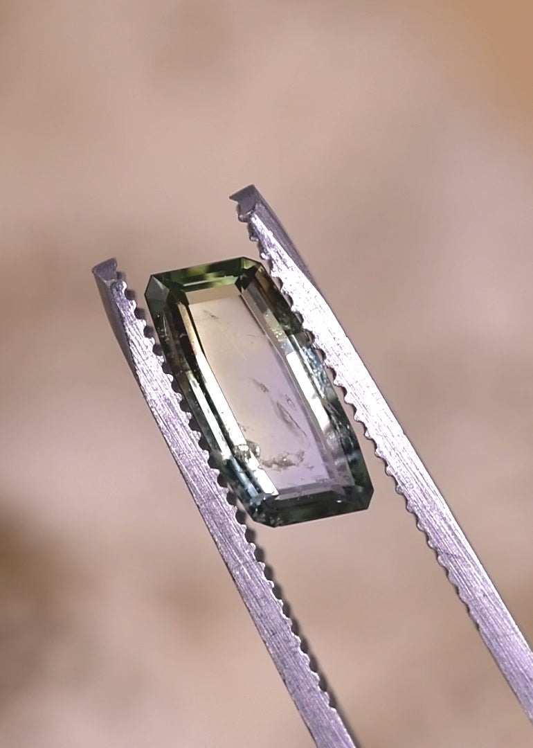 Close-up video of an unheated 2.31CT Umba River sapphire held in tweezers, showcasing elongated baguette portrait cut, crisp facet edges, and yellow green teal color play