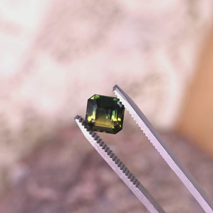 Rotating loose Australian parti sapphire held in tweezers, revealing the crisp Asscher cut facets and the interaction of green, yellow, and blue color zoning.