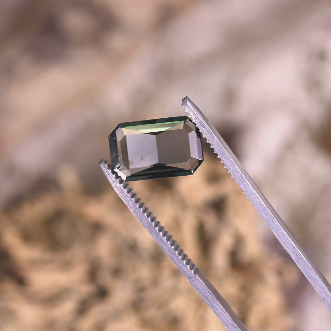 Close-up video of 2.96ct teal blue-green Tanzanite custom emerald cut held in tweezers, ultra low profile precision faceted gemstone from Tanzania displaying beveled corners and linear step brilliance