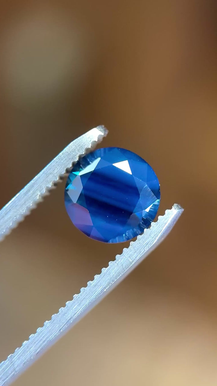 Round brilliant cut sapphire in tweezers displaying depth, saturation, and shifting opalescent effect.