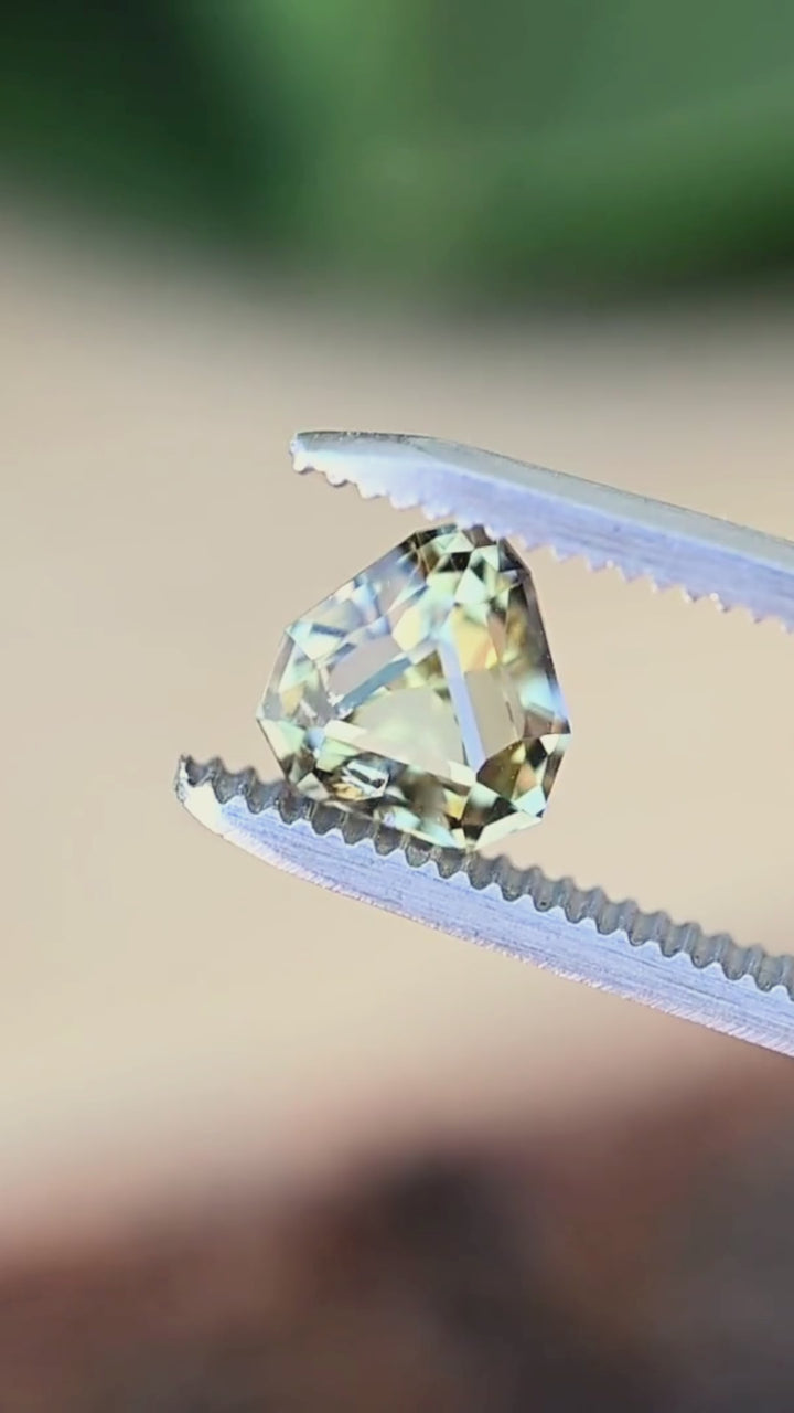 A custom geometric shield cut Tanzanite turning slowly in jeweler's tweezers, displaying crisp faceting, vibrant pleochroism, and dramatic flashes of teal, green, and yellow.