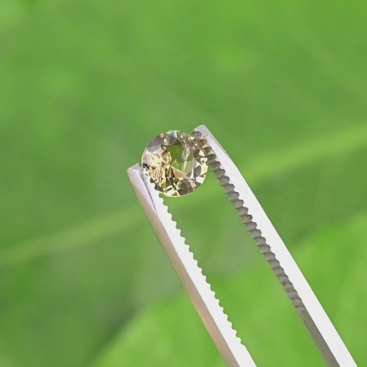1.04 carat golden orange Tanzanite round custom brilliant cut held in tweezers, slowly turned to highlight pavilion depth, crown facets, and natural Tanzania origin gemstone quality.