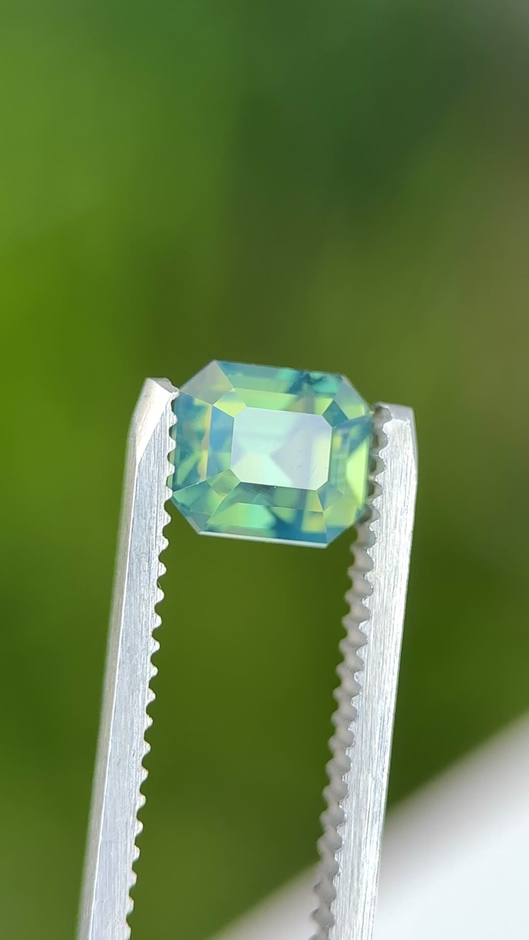 Precision emerald cut opalescent teal sapphire rotating in gem tweezers under natural light showing off luminous color and expert faceting.