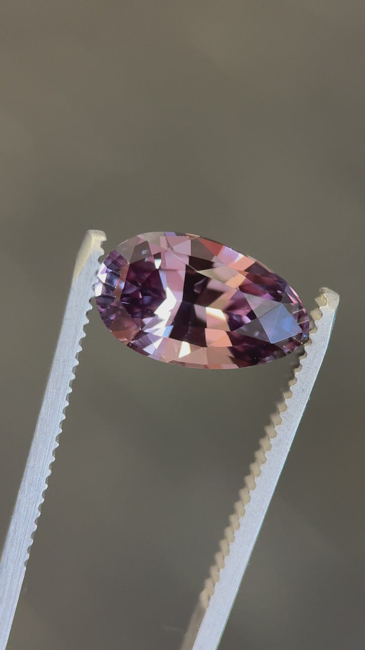 Loose heated color changing sapphire turning in tweezers, revealing purplish red and peach champagne tones in the modified brilliant pear cut.